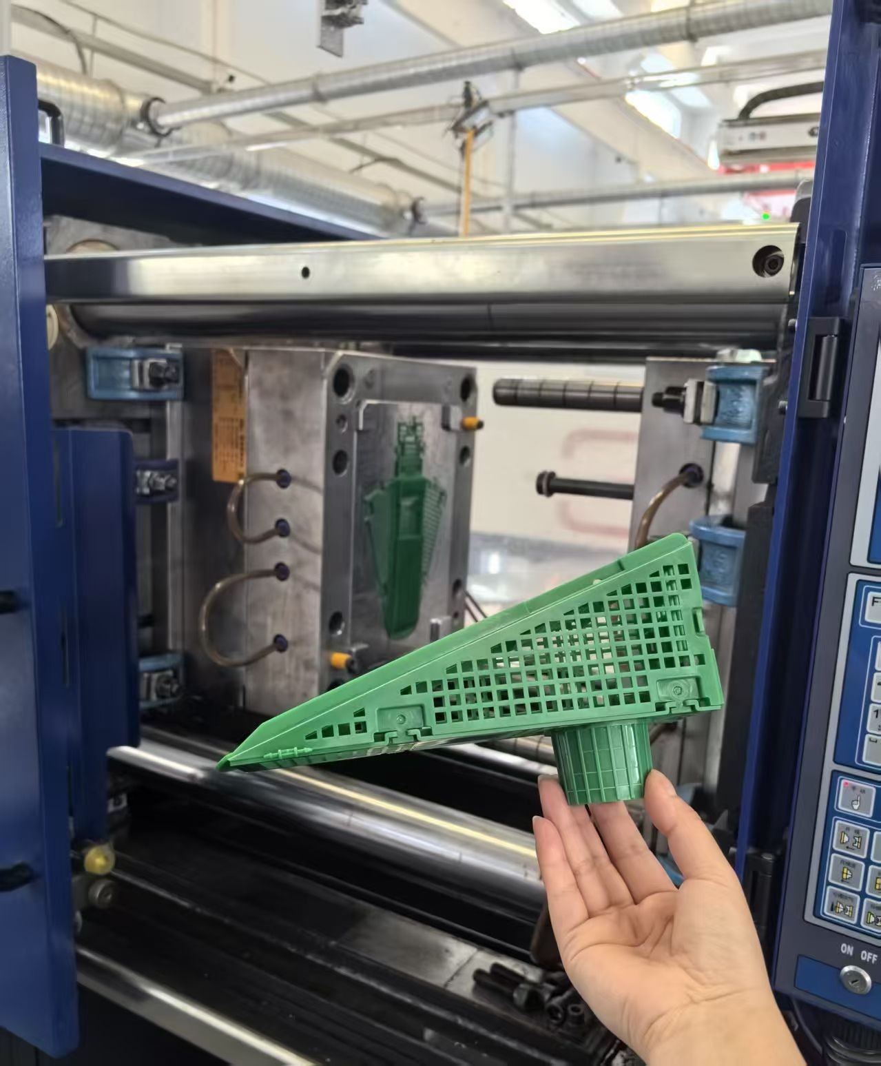 Hand holding green plastic part in front of an open mold inside an injection molding machine.