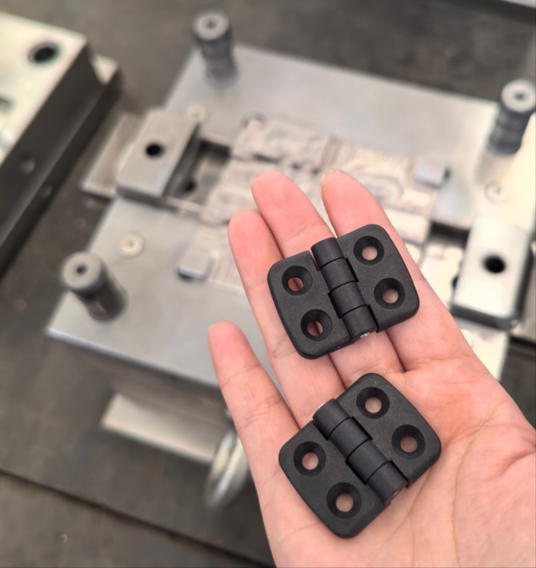 Hand holding two black plastic hinges, with a blurred industrial background.
