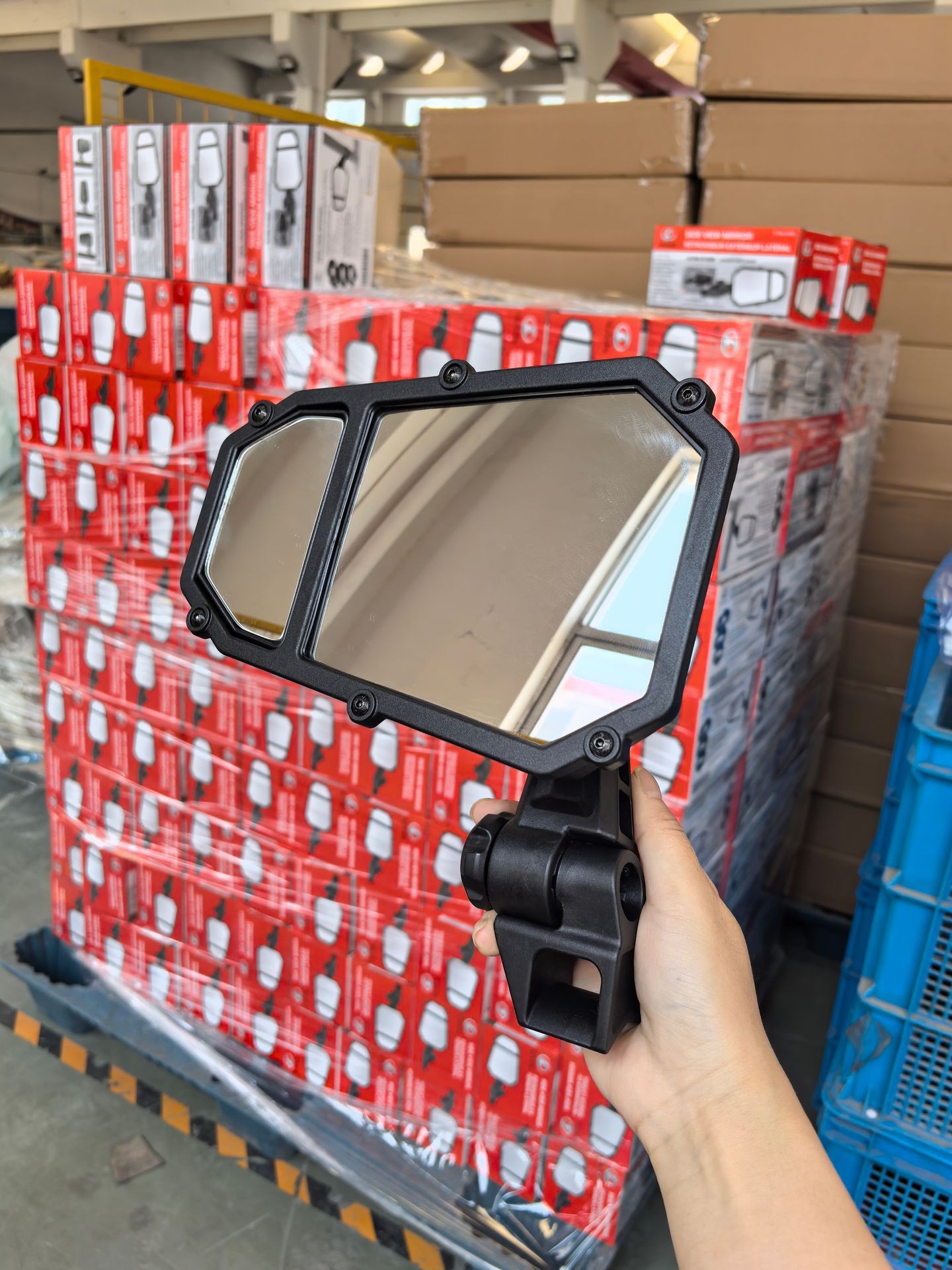 Person holding a side mirror in front of stacked boxes in a warehouse setting.