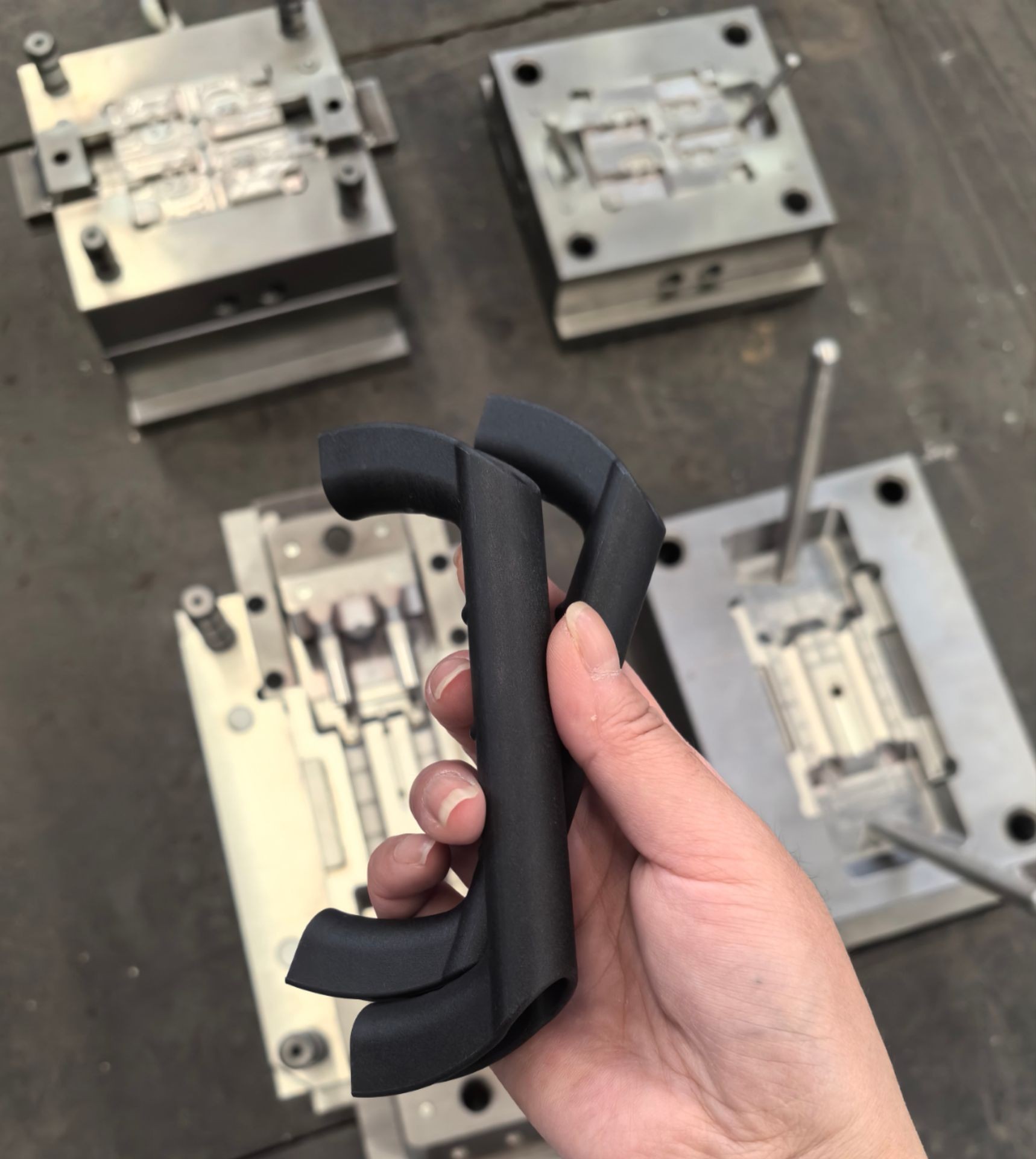 Hand holding black plastic parts with metal molds in the background on a workshop floor.